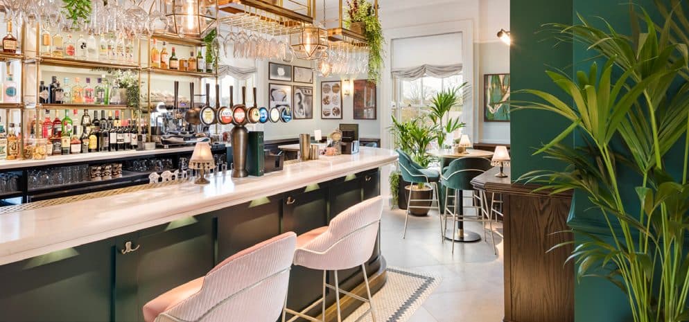 Bar at a Parogon Group restaurant, with brass shelving, pink velvet bar stools and potted palms